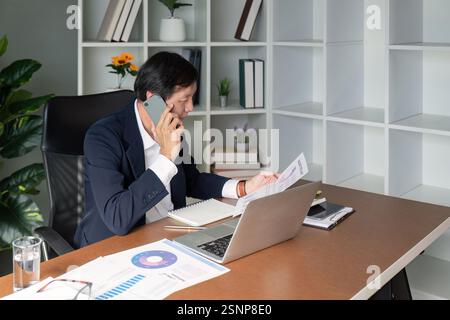 Elderly professional reviewing financial reports while on a call in a modern office environment. Stock Photo