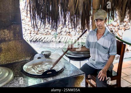 A cook stirs a large pan in an outdoor kitchen at a local coconut candy factory, Quoi Son, Vietnam . Monday, November 11, 2024. Stock Photo