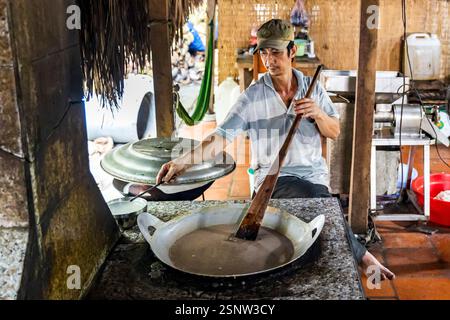 A cook stirs a large pan in an outdoor kitchen at a local coconut candy factory, Quoi Son, Vietnam . Monday, November 11, 2024. Stock Photo