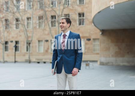 Businessman walking outside office holding laptop smiling Stock Photo