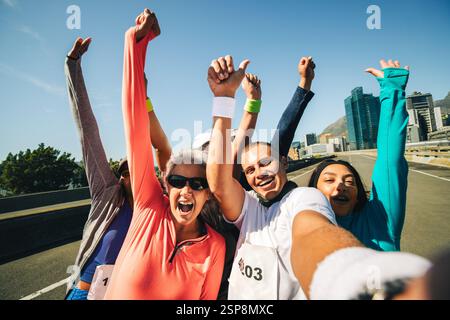 A group of diverse runners celebrates with raised arms after completing a race under a clear blue sky in the city. Their happy expressions and energet Stock Photo