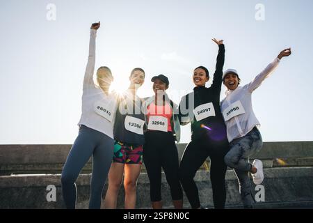 A group of diverse female friends celebrates afte a road race. They stand together with race bibs, arms raised victoriously, capturing the spirit of f Stock Photo