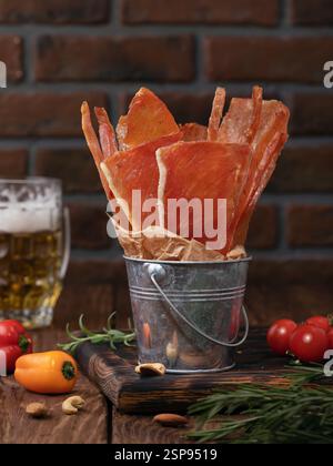 Smoked meat chips of different shapes from pork, chicken and turkey in a small decorative bucket on a wooden table with various vegetables, nuts and a Stock Photo
