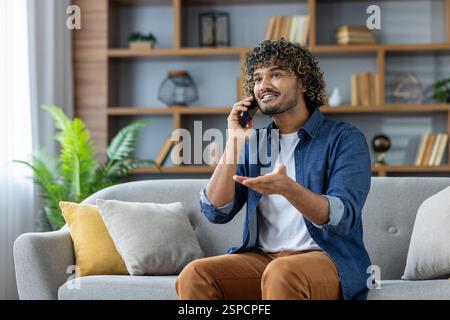 Young man talking on the phone while sitting in the living room on the sofa at home, smiling happily. Stock Photo