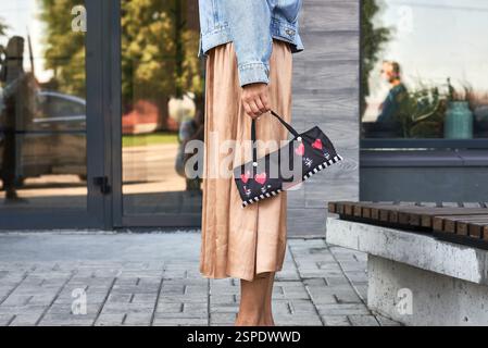 Stylishly, woman holds decorative purse with colorful hearts, dressed in denim jacket and skirt, urban street concept Stock Photo