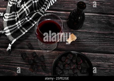 Red wine in a glass next to a black bottle and some blackberries on a wooden table in a dark moody rustic atmosphere, top view Stock Photo