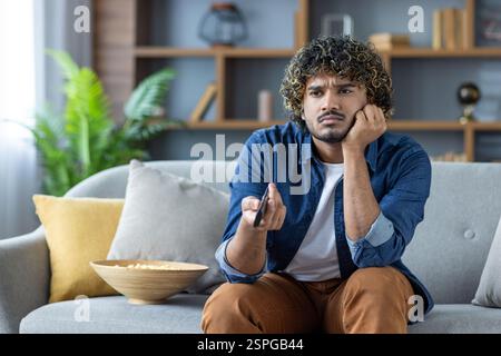 Bored and sad man sitting at home on the couch in the living room with the TV remote in his hands, depressed and unhappy alone at home. Stock Photo