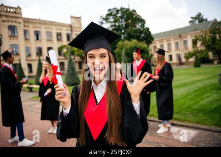 Joyful students celebrating graduation day outdoors on a sunny campus, wearing caps and gowns, holding diplomas. Stock Photo
