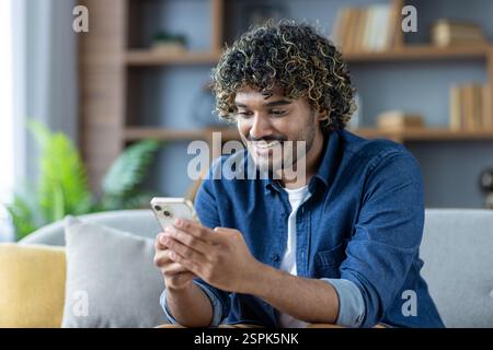 Young joyful man uses phone while sitting at home on sofa in living room of apartment, smiles happily, browses social media, and types text message. Stock Photo