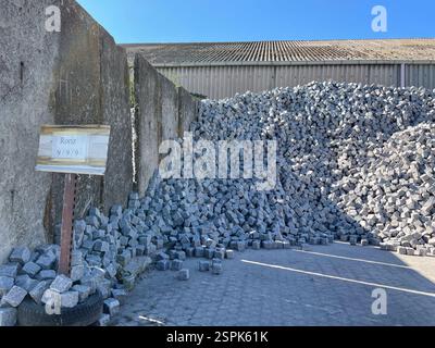 A large pile of light gray cubic paving stones sits against a concrete wall. A small sign indicates "Roriz 9/9".  The background shows a corrugated me Stock Photo