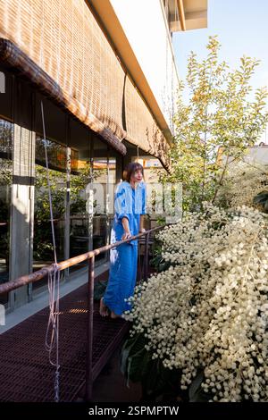 A barefoot woman in a relaxed blue outfit stands on a modern balcony, gently touching lush white blossoms. Surrounded by greenery and natural light, she enjoys a peaceful and refreshing moment outdoors Stock Photo