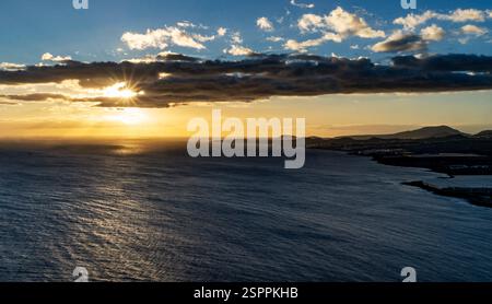 A stunning sunset over the ocean, with rays of sunlight breaking through clouds. The horizon features distant hills and a calm sea reflecting the gold Stock Photo