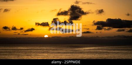 A serene sunset over the ocean, with the sun partially hidden behind clouds. The sky is painted in shades of orange and yellow, reflecting on the calm Stock Photo