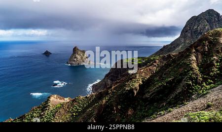 A breathtaking coastal landscape featuring rugged mountains, a rocky shoreline, and a distant island. The sky is partly cloudy with hints of sunlight Stock Photo