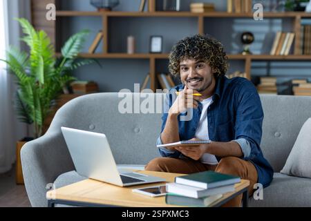 Portrait of young student studying remotely at home. Smiling man sitting on sofa in living room, using laptop for video call. Stock Photo
