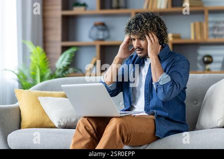 Upset man sitting on sofa in living room at home, in apartment. Employee working remotely using laptop, received notification, error message. Stock Photo