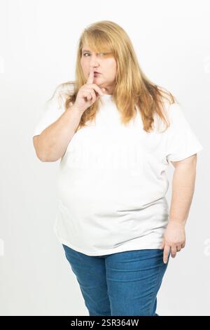 Portrait of a blonde plus size woman with her finger on her lips, signaling silence. Stock Photo