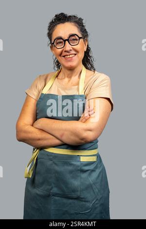 Confident mature woman in apron and glasses crossing arms, exuding professionalism and expertise in culinary or domestic setting Stock Photo