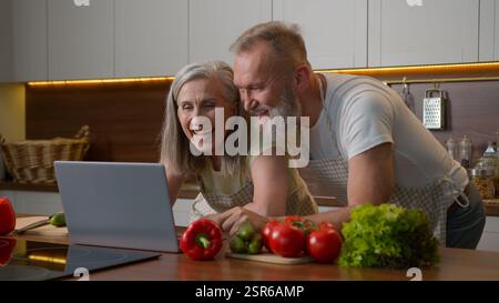 Older Caucasian couple middle-aged spouses family using laptop device at home kitchen choosing food delivery fresh vegetables online shopping natural Stock Photo