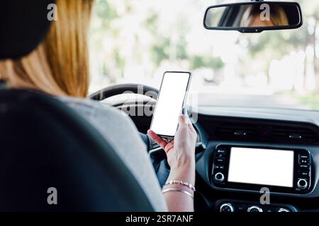 Female driver interacts with her mobile phone and the car's digital control screen. Blank display of smartphone and car computer screen for mockup. Stock Photo