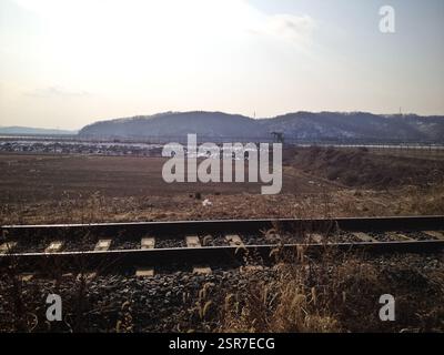 Railway tracks stretch across a vast, open field, bordered by distant hills under a pale sky. The tracks, once bustling with trains, now stand solitar Stock Photo