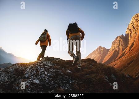 Silhouettes of two active sporty young hikers walks with backpacks high in mountains against sunset sky Stock Photo
