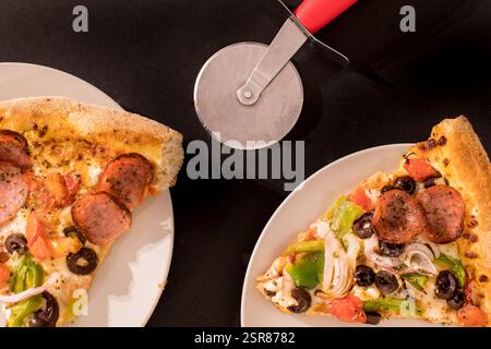 Two Slices of Different Pizzas with Pepperoni, Olives, and Vegetables, Beside a Pizza Cutter on a Dark Background Stock Photo