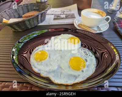 Three fried eggs on a shiny plate, a basket of bread and a cappuccino as breakfast Stock Photo