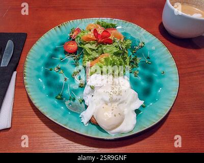 Poached egg with fresh vegetables (greens, lettuce, tomato, cress) and a decorative red edible flower on a plate and cappuccino as breakfast or lunch Stock Photo