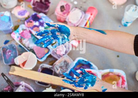Adult hands covered in vivid blue paint, with scattered art supplies outdoors. Soft lighting casts gentle shadows on ground, enhancing artistic mood Stock Photo