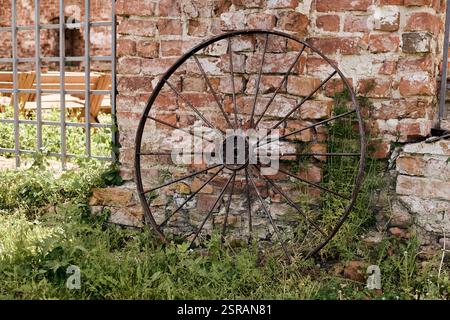 Old rusty metal wagon wheel resting against weathered brick wall, surrounded by overgrown grass and foliage Stock Photo