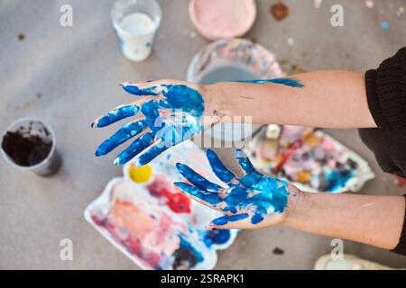 Adult hands covered in vivid blue paint, with scattered art supplies outdoors. Soft lighting casts gentle shadows on ground, enhancing artistic mood Stock Photo