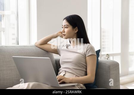 Woman gazing thoughtfully into distance seated on sofa with laptop Stock Photo