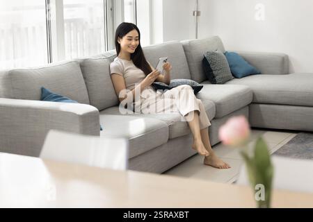 Pretty relaxed Asian single woman using smartphone seated on couch Stock Photo