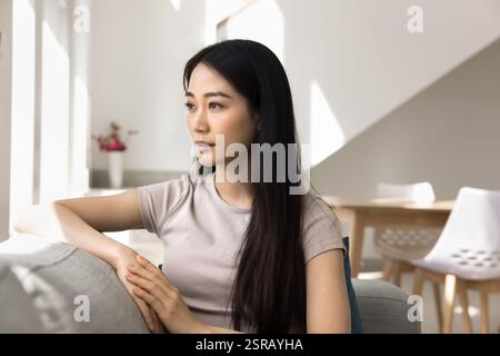 Serious Asian young woman sits thoughtfully on comfortable sofa Stock Photo