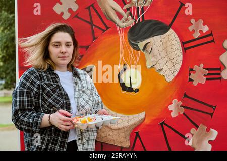 Young woman holding paint palette stands by vibrant surreal clock painting with puzzle patterns. Outdoor scene filled with creativity and orange hues Stock Photo