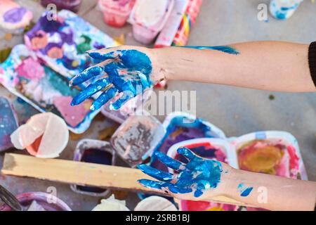 Adult hands covered in vivid blue paint, with scattered art supplies outdoors. Soft lighting casts gentle shadows on ground, enhancing artistic mood Stock Photo