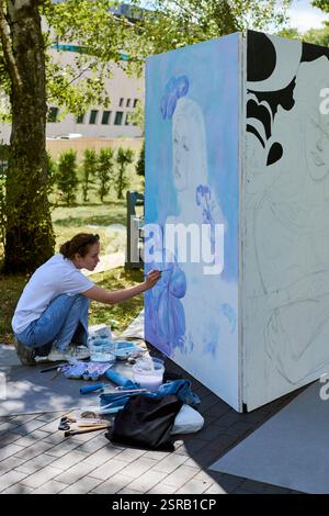 Young caucasian female artist, focused and engaged, paints mural outdoors on sunny day. Blue and white hues dominate artwork. Background features tree Stock Photo