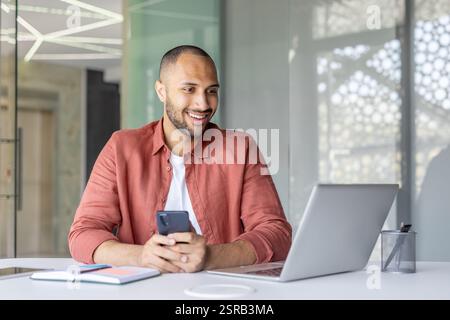 Successful joyful man with phone in hands smiling working inside office with laptop. Company employee satisfied with results of achievement at workplace. Stock Photo