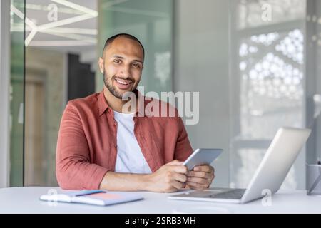 Portrait of a young successful man office worker programmer developer, looking at the camera smiling, with a tablet computer in his hands. Sitting at a desk with a laptop. Stock Photo
