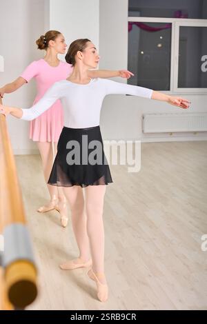 Two young female ballerinas with brown hair rehearsing at ballet barre. Wearing leotards and skirts, focused on technique. Bright studio with wooden f Stock Photo