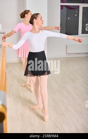Two young female ballerinas with brown hair rehearsing at ballet barre. Wearing leotards and skirts, focused on technique. Bright studio with wooden f Stock Photo