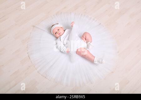 Infant female with light hair, lying on wooden floor in full white tutu. Expression calm and curious. Soft lighting enhances serene setting Stock Photo