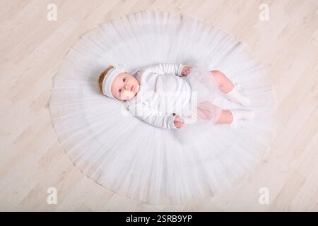 Infant female with light hair, lying on wooden floor in full white tutu. Expression calm and curious. Soft lighting enhances serene setting Stock Photo