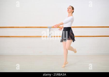 Young female ballerina with brown hair practicing in dance studio. Standing on pointe with focused expression. Bright lighting against simple, white b Stock Photo