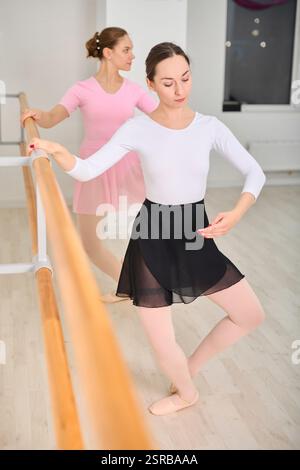 Two young female ballerinas practice at ballet barre in bright studio. One wears black skirt and white top other, pink dress. Light tones create seren Stock Photo