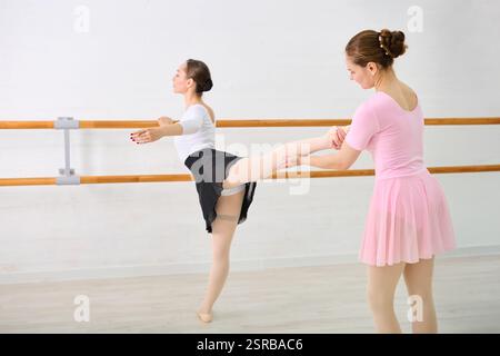 Two young female dancers with brown hair practice at ballet barre. One balances in arabesque, tutored by another wearing pink. Bright room enhances fo Stock Photo