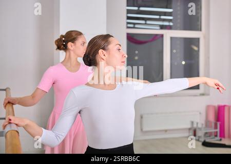 Two young white female ballerinas with light brown hair elegantly practice at barre in ballet studio. Pink and white attire contrasts softly with brig Stock Photo