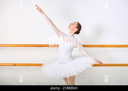 Young female ballerina with light brown hair practicing elegant pose at ballet barre. White leotard and tutu enhance serene expression against soft-li Stock Photo
