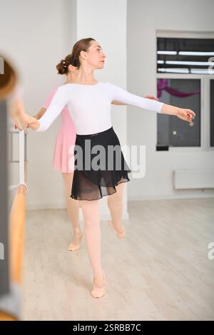 Two young female ballerinas practice at ballet barre in bright studio. One wears black skirt and white top other, pink dress. Light tones create seren Stock Photo